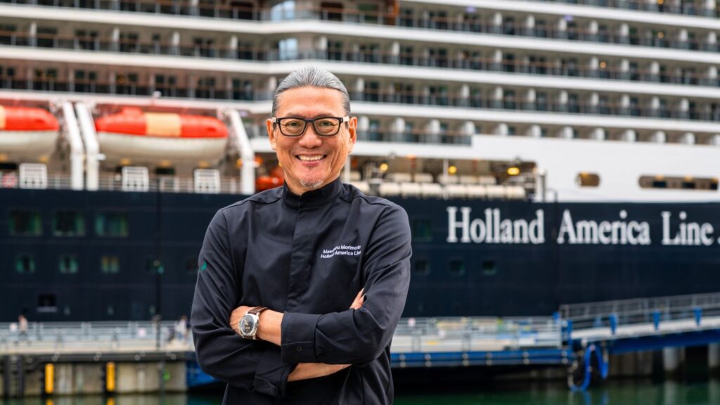 A man wearing glasses and a black chef&rsquo;s coat stands with arms crossed in front of a Holland America Line cruise ship docked at a port.
