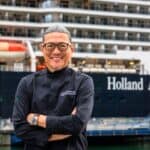 A man wearing glasses and a black chef’s coat stands with arms crossed in front of a Holland America Line cruise ship docked at a port.