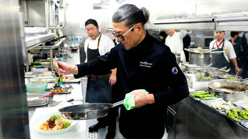 A chef in a black uniform prepares a dish in a busy restaurant kitchen, surrounded by other kitchen staff and various cooking utensils.