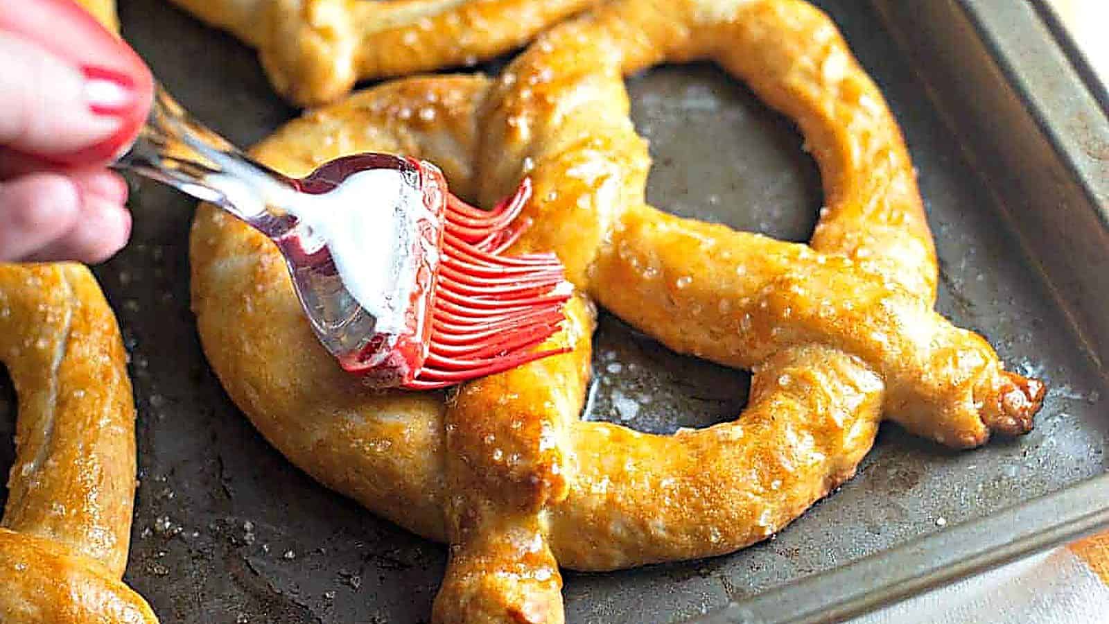 A hand uses a red silicone brush to apply butter or glaze to a baked soft pretzel on a baking sheet.