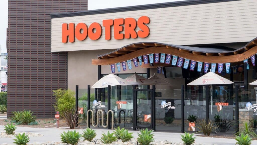 Exterior of a Hooters restaurant with orange signage, outdoor seating with umbrellas, decorative plants, and hanging pennant banners above the entrance&mdash;a lively scene from one of the restaurant chains that came back and regained its place in popular dining culture.