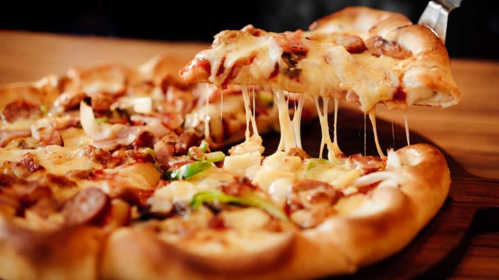 A close-up of a pizza with melted cheese, vegetables, and meat. A slice is being lifted, showing stretchy cheese.