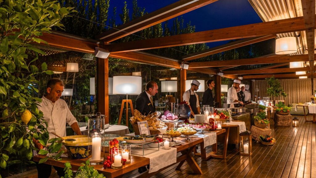 Chefs stand behind buffet tables with various dishes, candles, and fresh produce at an outdoor evening event under a wooden pergola.