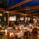 Chefs stand behind buffet tables with various dishes, candles, and fresh produce at an outdoor evening event under a wooden pergola.