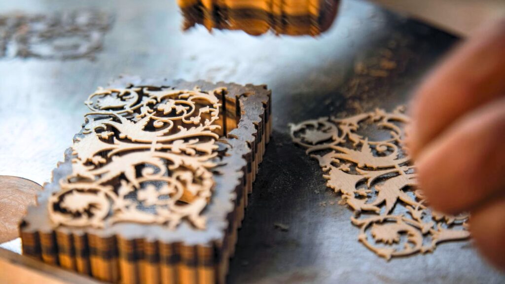 Close-up of a decorative wooden piece being laser cut, showing intricate patterns and a hand reaching toward the finished cutout.
