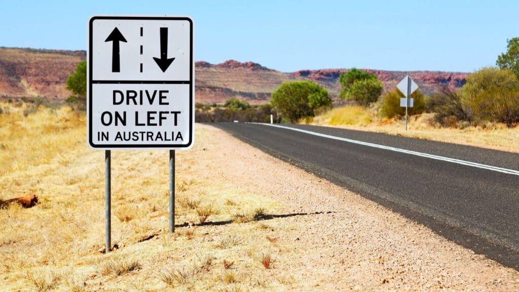 Roadside sign in Australia reads &ldquo;DRIVE ON LEFT IN AUSTRALIA,&rdquo; with arrows indicating driving directions&mdash;a helpful reminder for visitors from countries driving on the opposite side; dry landscape and empty road in background.