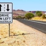 Roadside sign in Australia reads “DRIVE ON LEFT IN AUSTRALIA,” with arrows indicating driving directions—a helpful reminder for visitors from countries driving on the opposite side; dry landscape and empty road in background.