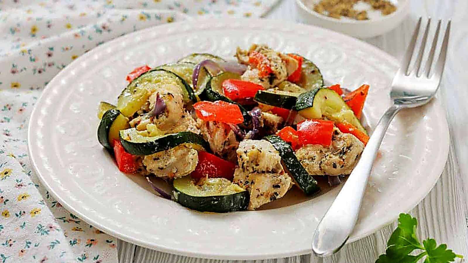 A white plate with a serving of Italian Herbed Chicken & baked foil packs with a fork on the side and a small bowl of seasoning in the background.