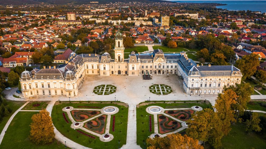 Aerial view of a large historic palace with symmetrical gardens in the foreground, surrounded by a town and distant hills; reminiscent of the lush landscapes you&rsquo;ll find when exploring Hungary during the best time to visit Lake Balaton.