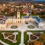 Aerial view of a large historic palace with symmetrical gardens in the foreground, surrounded by a town and distant hills; reminiscent of the lush landscapes you’ll find when exploring Hungary during the best time to visit Lake Balaton.