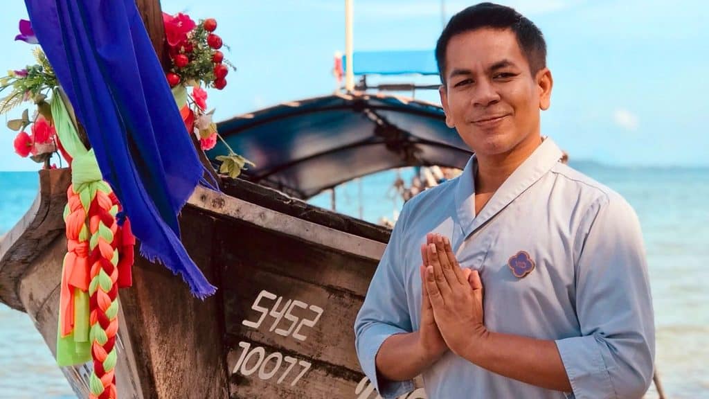A man in traditional attire stands by a decorated wooden boat on a beach at Phulay Bay Krabi, hands pressed together in greeting, with the sea and sky stretching beautifully in the background.