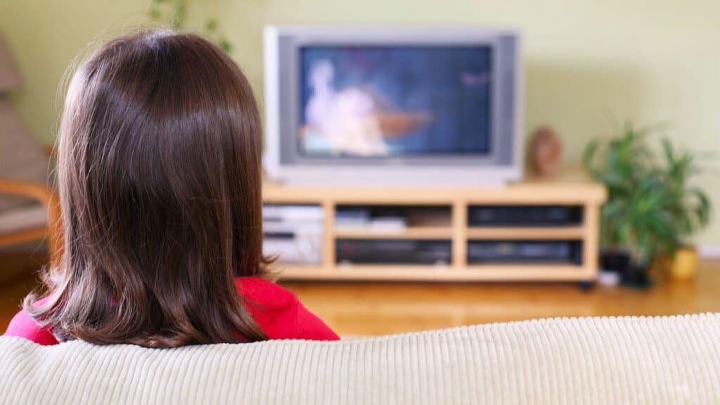 A child with brown hair sits on a couch facing a television in a living room with a wooden media console and green plants, enjoying the familiar comfort of Saturday cartoon traditions.