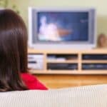 A child with brown hair sits on a couch facing a television in a living room with a wooden media console and green plants, enjoying the familiar comfort of Saturday cartoon traditions.