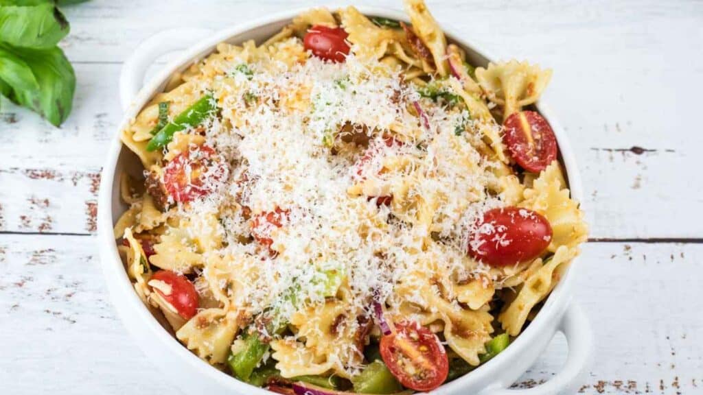 Bowl of lasagna pasta salad with cherry tomatoes, green beans, and grated cheese on top, placed on a white wooden surface.