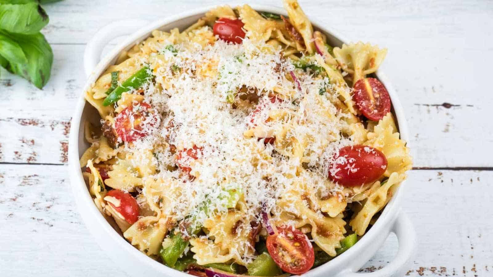 Bowl of lasagna pasta salad with cherry tomatoes, green beans, and grated cheese on top, placed on a white wooden surface.