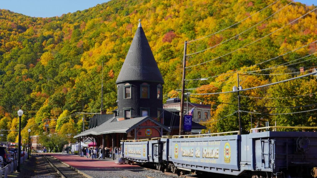 A historic train station with a pointed tower sits beside a blue train; wooded hills with autumn foliage rise in the background.