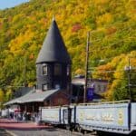 A historic train station with a pointed tower sits beside a blue train; wooded hills with autumn foliage rise in the background.