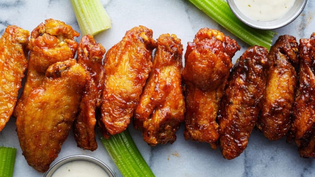 A row of glazed chicken wings is arranged on a marble surface with celery sticks and small cups of ranch dressing, offering a delicious nod to classic Buffalo wings facts and traditions.
