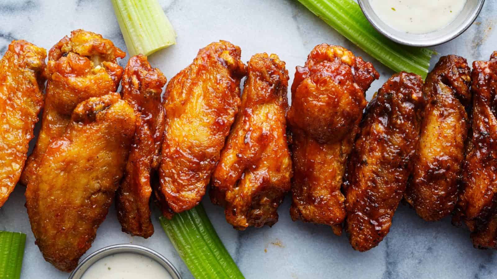 A row of glazed chicken wings is arranged on a marble surface with celery sticks and small cups of ranch dressing, offering a delicious nod to classic Buffalo wings facts and traditions.