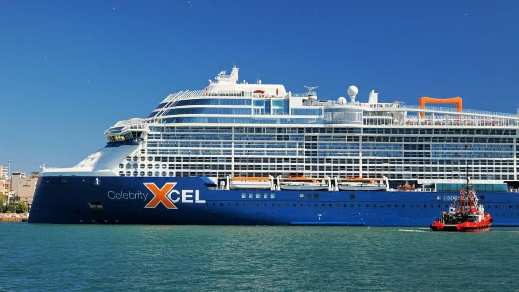 Large blue and white cruise ship labeled &ldquo;Celebrity Xcel&rdquo; docked near shore, with a small red tugboat nearby on calm water under a clear blue sky.
