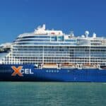 Large blue and white cruise ship labeled “Celebrity Xcel” docked near shore, with a small red tugboat nearby on calm water under a clear blue sky.