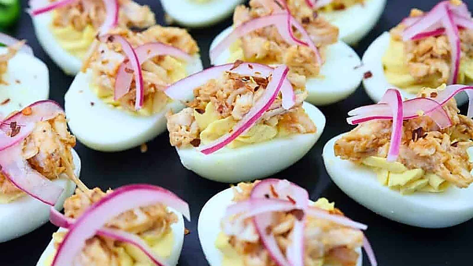 Deviled eggs topped with shredded meat and thinly sliced red onions are arranged on a black plate.