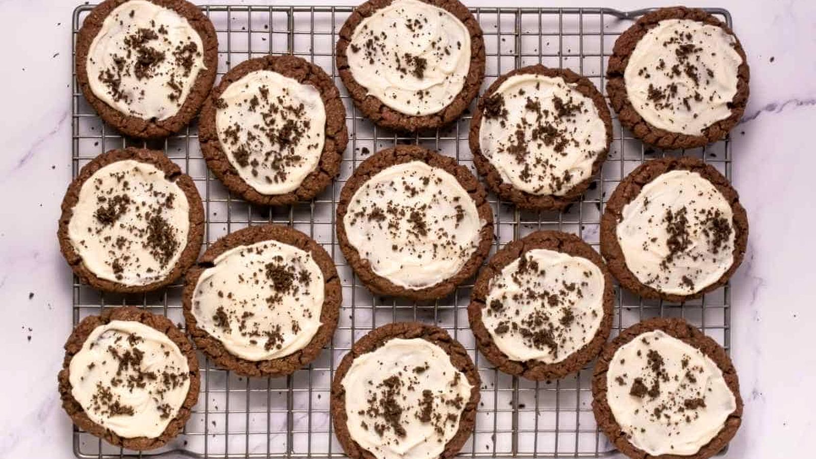 A cooling rack with twelve Crumbl Copycat Frosted Oreo Cookies topped with white frosting and chocolate crumbs, arranged on a white marble surface.