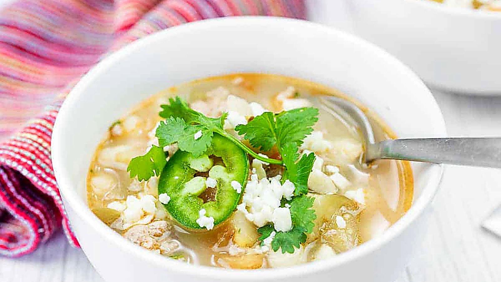 A bowl of Green pork posole soup garnished with cilantro, sliced jalape&ntilde;o, and crumbled cheese, with a spoon resting inside and a striped cloth in the background.