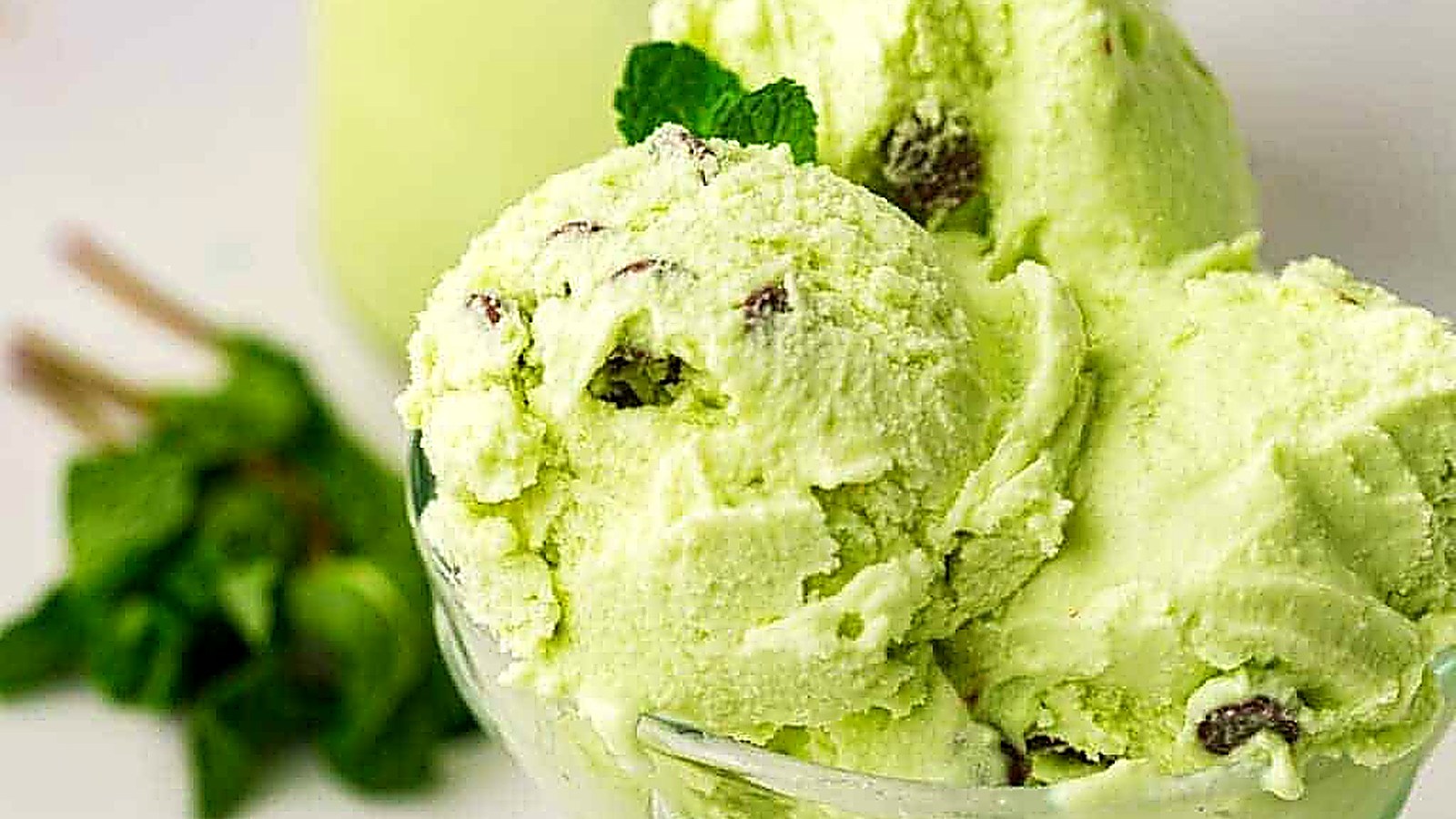 Three scoops of mint chocolate chip ice cream in a glass bowl, with chocolate pieces visible and a sprig of mint in the background.