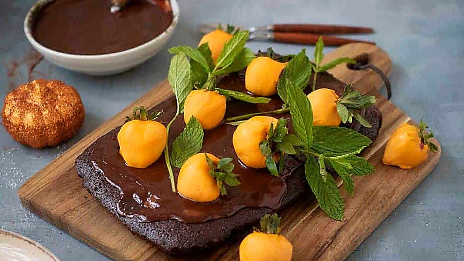 A rectangular Halloween Pumpkin Patch Brownie Cake, topped with yellow fruit resembling mini pumpkins and fresh mint, sits on a wooden board.