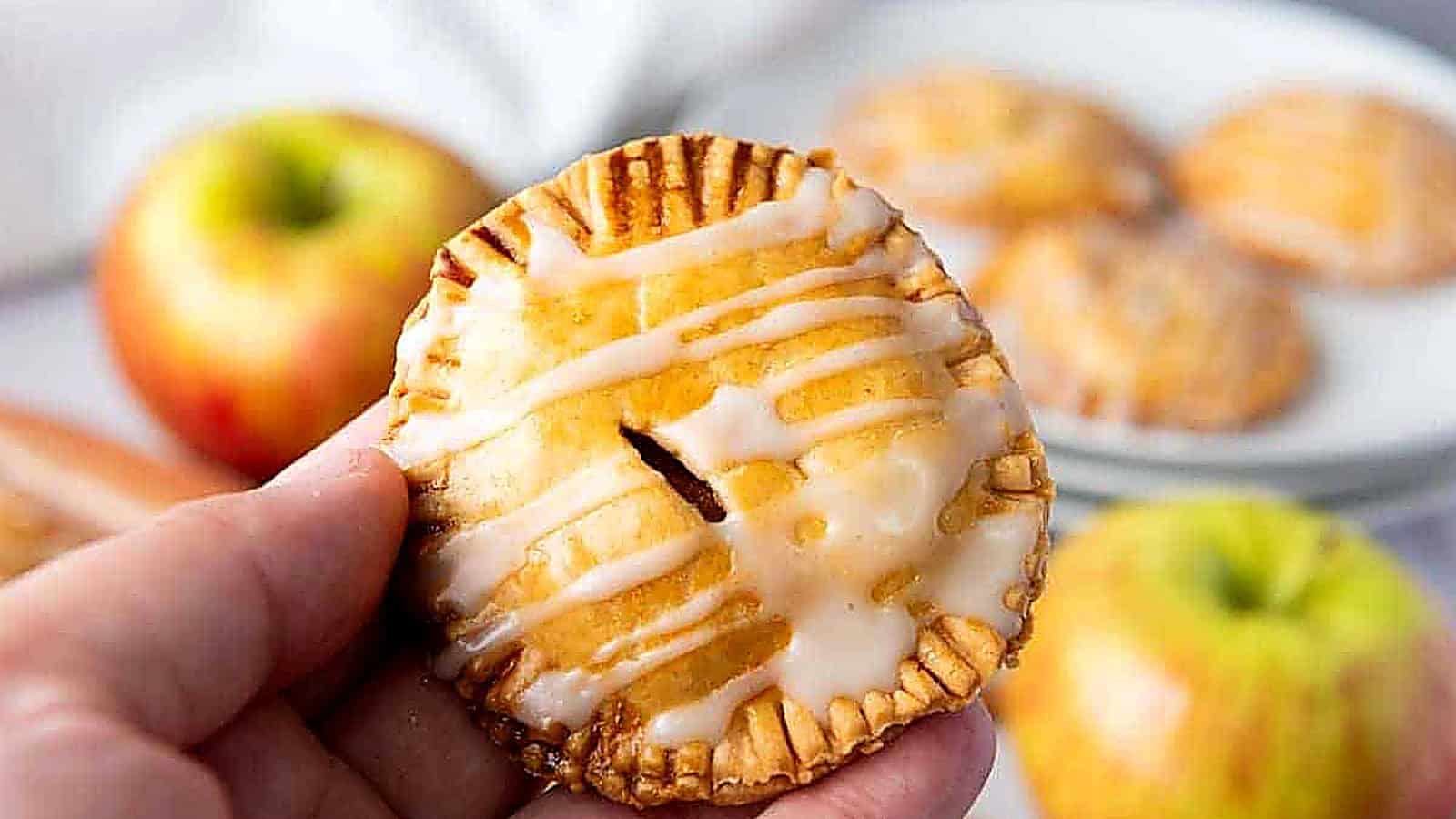 A hand holds a small round hand pie with icing drizzle; apples and more hand pies are visible in the background.