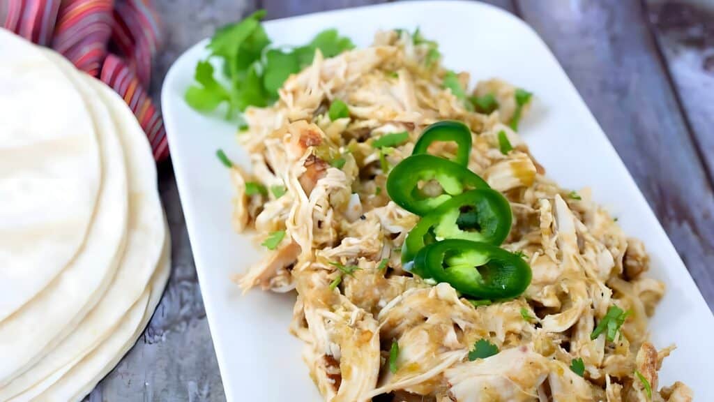 Shredded chicken topped with sliced jalape&ntilde;os and cilantro on a white plate, with tortillas and a striped cloth napkin beside it.