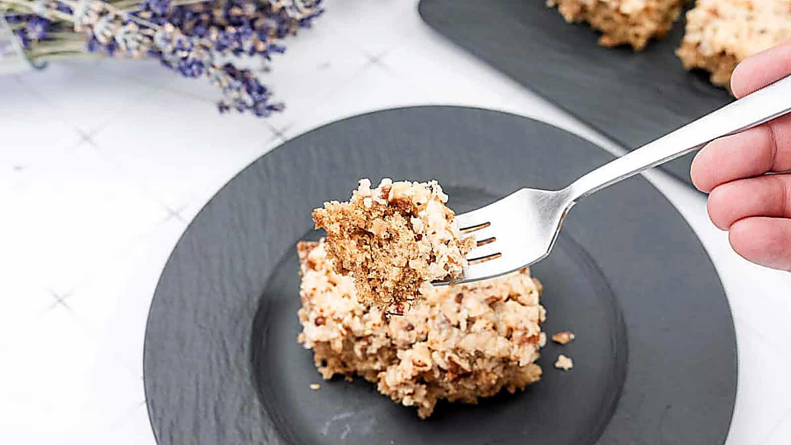 A fork holds a bite of Old Fashioned Lazy Daisy Oatmeal Cake above a black plate with more cake pieces, with dried lavender in the background.