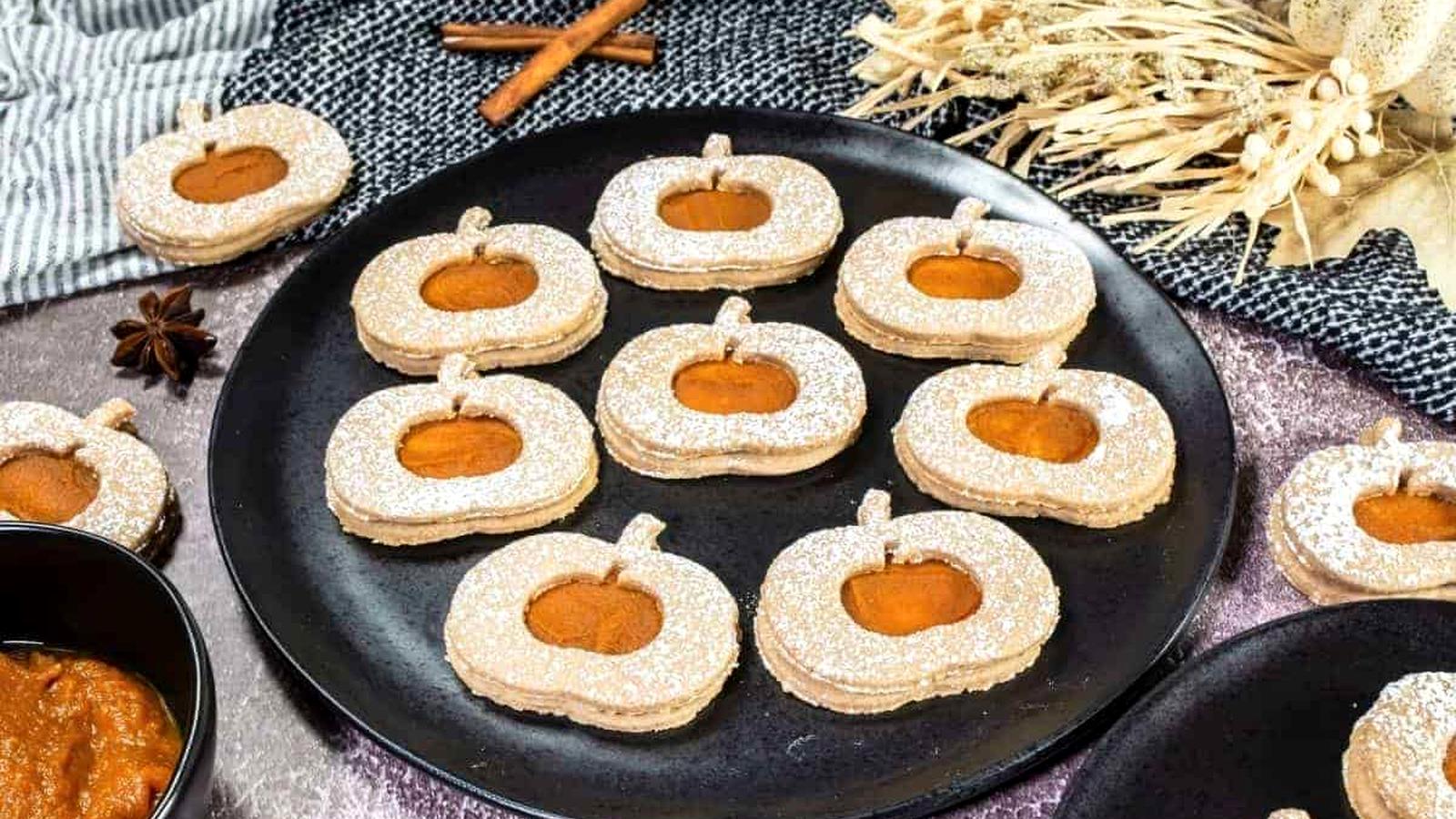 A black plate holds Pumpkin Linzer Cookies with powdered sugar and pumpkin filling. Other cookies and a bowl of filling are nearby on a textured tablecloth.