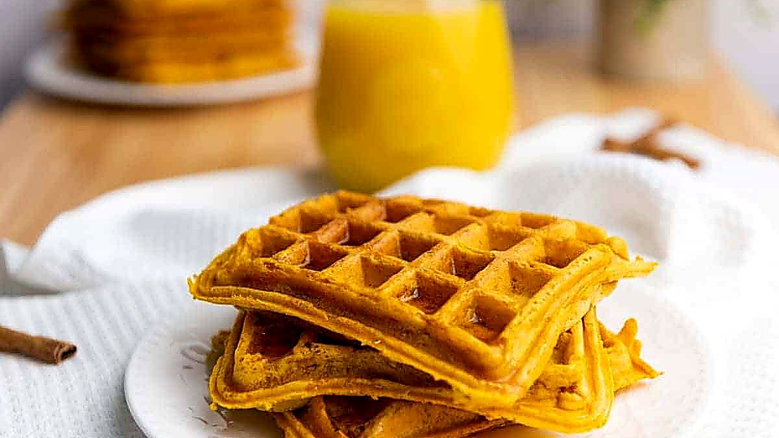 A plate with three stacked pumpkin waffles drizzled with syrup, with a glass of orange juice and more waffles in the background.