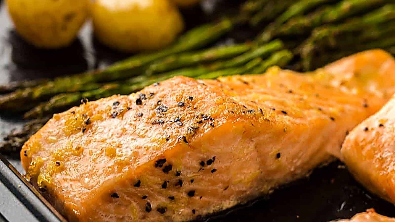 Close-up of a baked salmon fillet seasoned with pepper, served alongside asparagus spears and small roasted potatoes.
