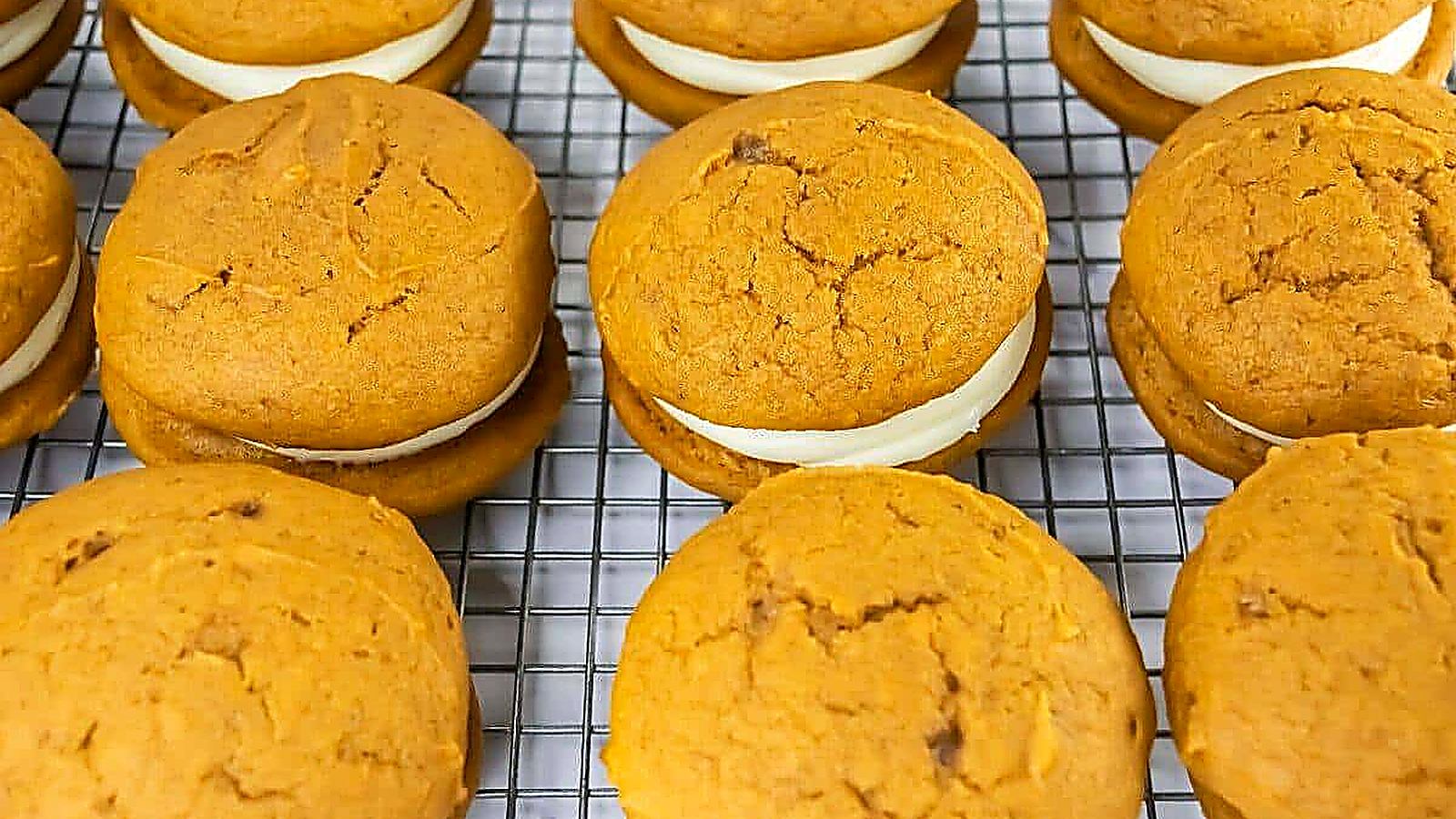 Pumpkin whoopie pies with cream filling are arranged in rows on a wire cooling rack.