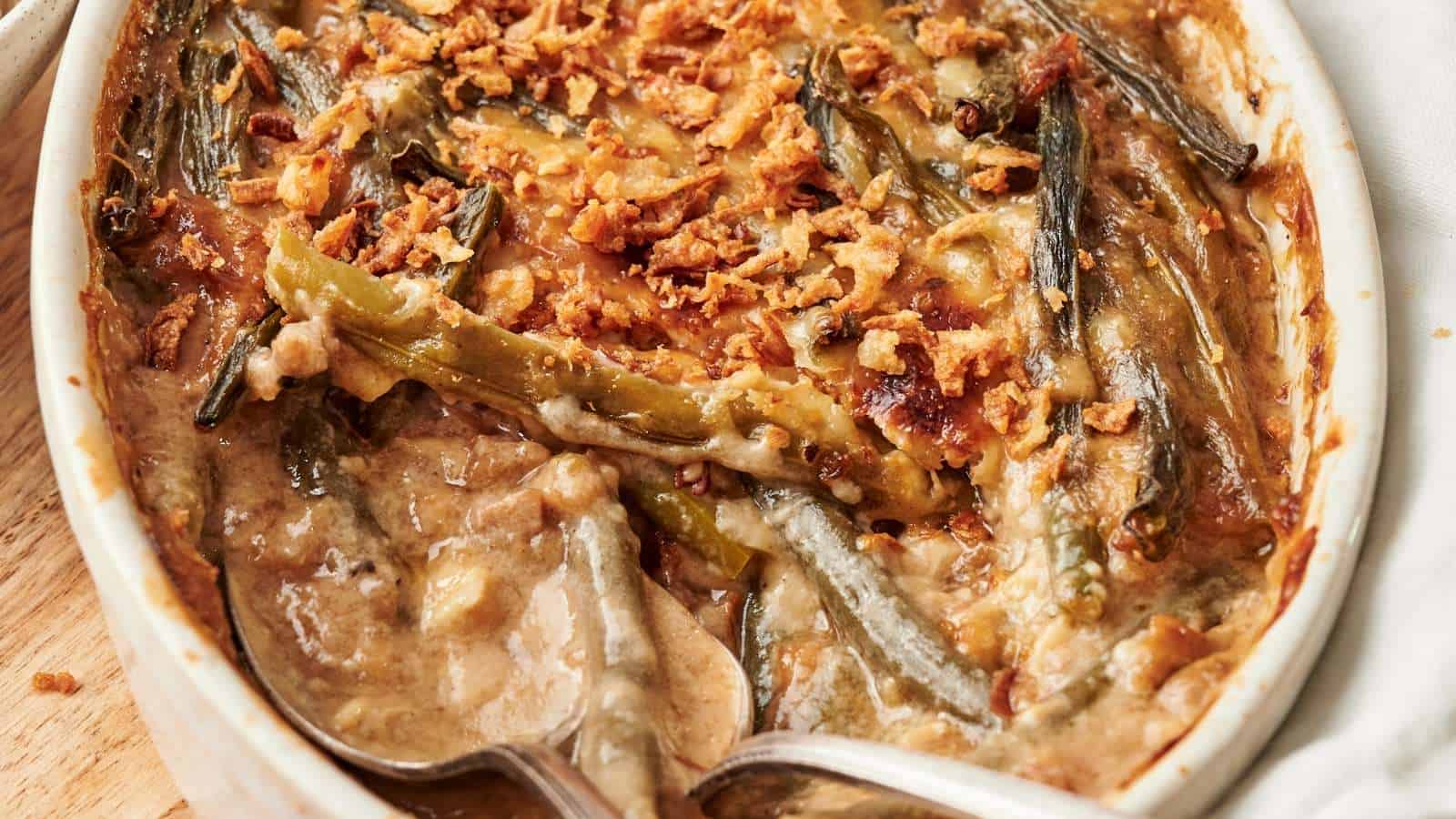 A baked green bean casserole topped with crispy fried onions, partially served with a metal spoon in a white oval dish.