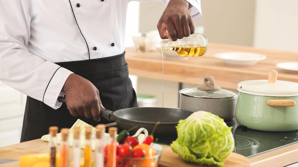 A chef pours cooking oil into a frying pan on a stove, with fresh vegetables, a pot, and kitchen utensils visible on the countertop.