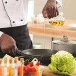 A chef pours cooking oil into a frying pan on a stove, with fresh vegetables, a pot, and kitchen utensils visible on the countertop.