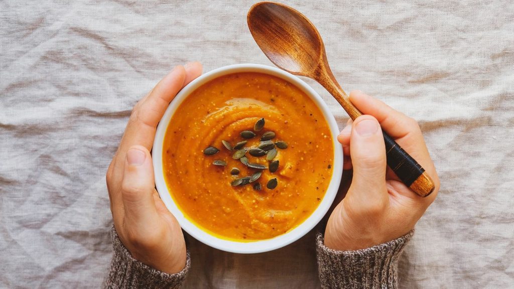Person holding a bowl of orange soup topped with pumpkin seeds, with a wooden spoon in one hand, on a beige cloth background—a simple scene that captures why we crave comfort food.