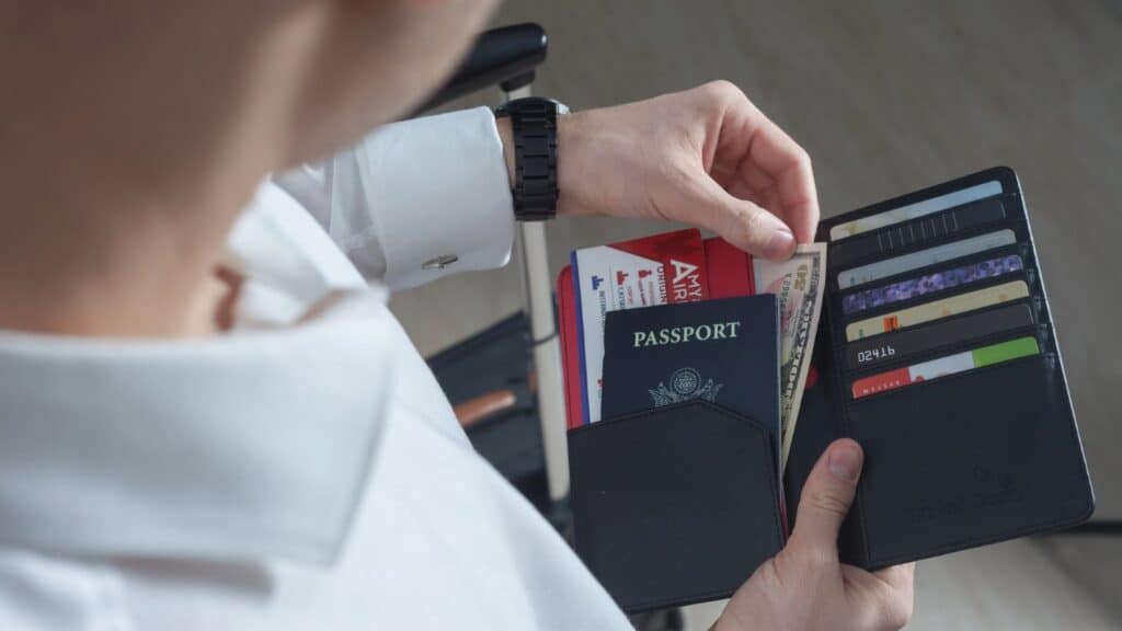 A person holding a travel wallet containing a passport, airline boarding pass, and U.S. currency to ensure they are prepared in case a debit card is declined abroad.