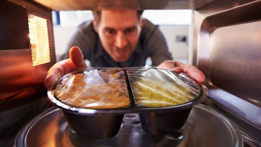 A man places a plastic-sealed microwave meal inside a microwave oven, with the meal centered on the glass turntable.