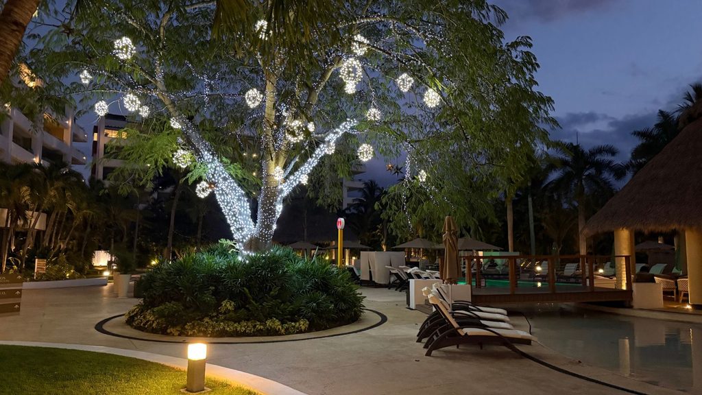 A poolside area at dusk at Marival Distinct All-Suites & World Spa features a large tree decorated with white string lights and hanging ornaments, surrounded by lounge chairs, lush greenery, and elegant resort buildings.