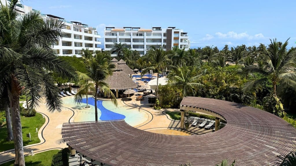 Resort pool area with palm trees, lounge chairs, thatched roofs, and white modern buildings in the background under a clear blue sky.