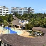 Resort pool area with palm trees, lounge chairs, thatched roofs, and white modern buildings in the background under a clear blue sky.