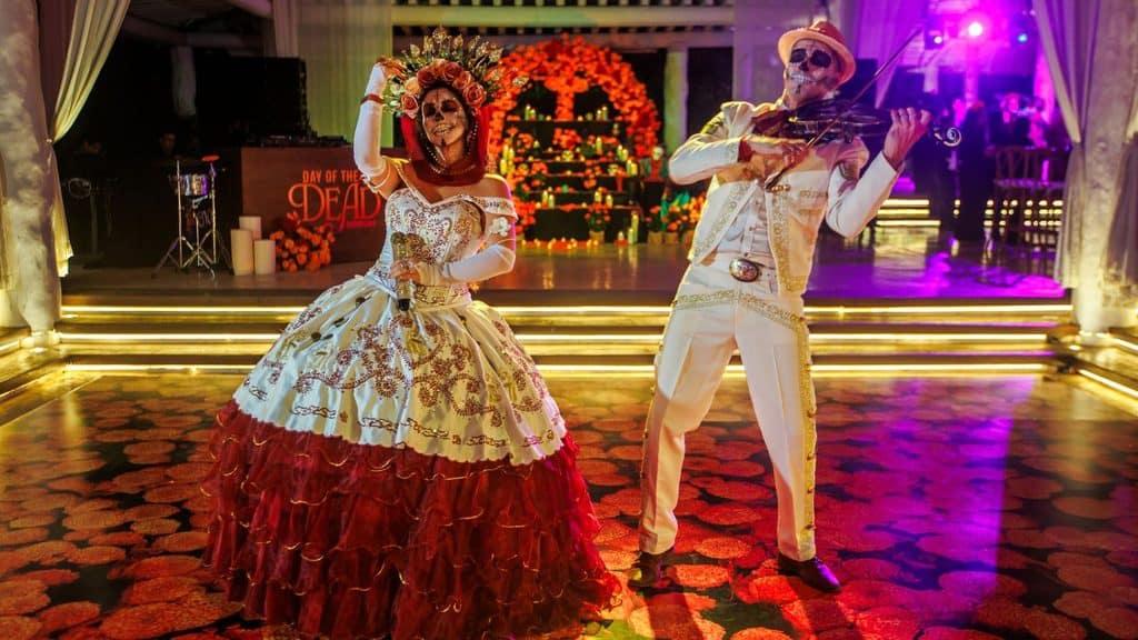 Two people dressed in elaborate Day of the Dead costumes and face paint perform with a violin in a beautifully decorated venue in Mayakoba, with marigold petals scattered across the floor.