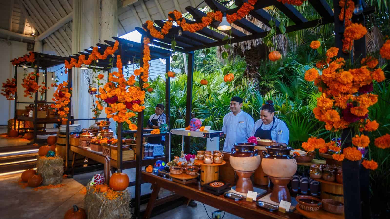 Buffet tables decorated with orange marigolds and pumpkins evoke a festive ambiance at Mayakoba. Chefs stand behind the tables serving food, while lush greenery provides a beautiful backdrop.