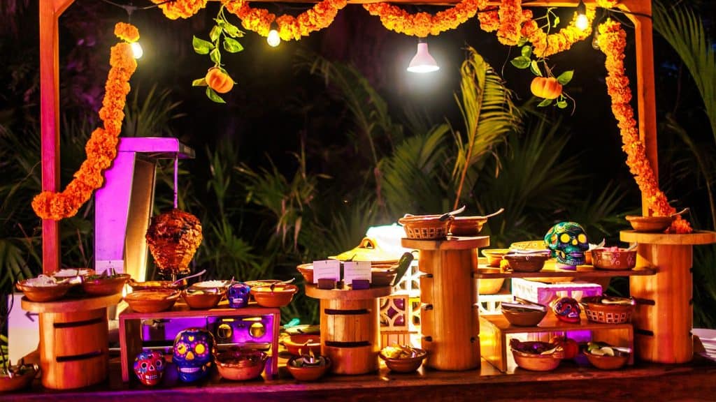Wooden food stand with various bowls of food, decorated with marigold garlands and colorful skulls, illuminated by hanging lights. Lush greenery in the background evokes the vibrant ambiance of Mayakoba.