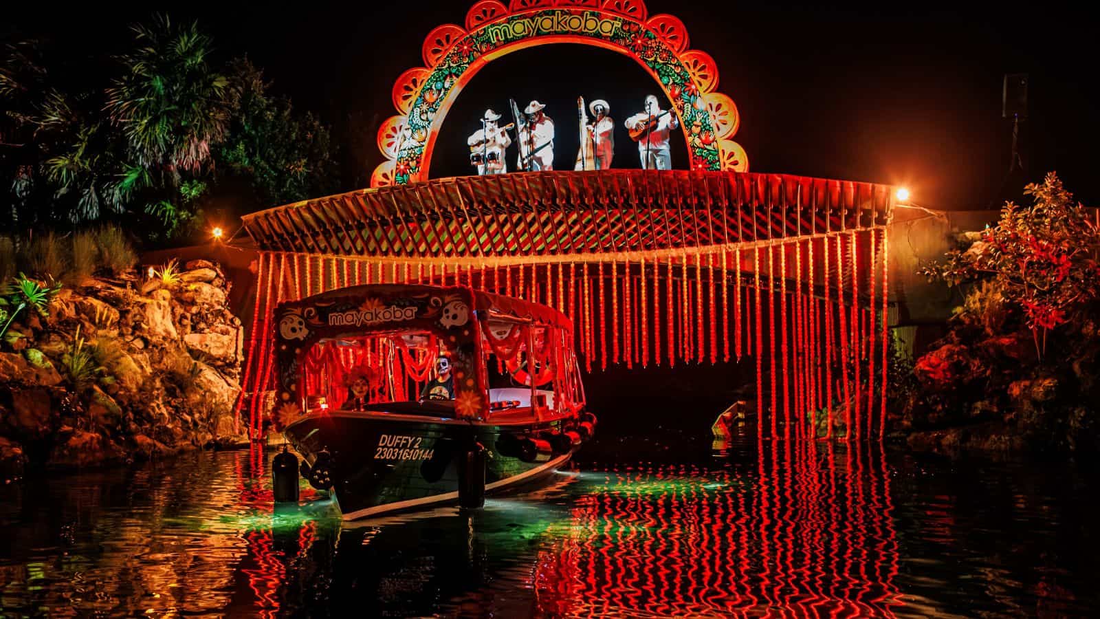 A boat with red lights passes under a decorated bridge at night in Mayakoba, with performers standing above the bridge under colorful arch lighting.